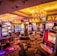 A vibrant casino interior filled with rows of colorful slot machines, each illuminated with bright lights and displays. The ceiling is adorned with elegant chandeliers and golden drapes, creating a luxurious atmosphere. The patterned carpet adds to the elaborate design of the space.