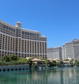 A large luxury hotel and casino building with multiple stories, featuring a light facade with arched windows. The structure has a prominent central tower with a dome-like top. It is surrounded by landscaping with trees and a body of water in the foreground.