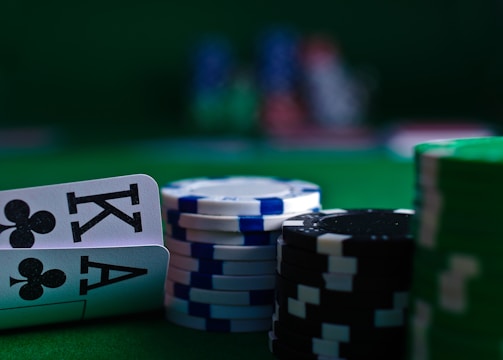 A casino scene featuring two playing cards, the ace and king of clubs, partially concealed by a player's hand. Nearby, there are stacks of multicolored poker chips on a green felt table, with blurred colorful background elements suggesting other chip stacks or cards.