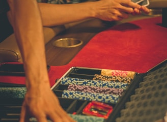 A person reaches into a poker chip case on a red felt table, while another person holds playing cards. The scene is dimly lit, creating a warm and engaged atmosphere.