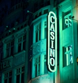 A vibrant neon sign displaying the word 'CASINO' is prominently illuminated against the facade of a building, showcasing intricate architectural details and a classic design. The sign emits a bright teal glow, casting light on the surrounding area and creating a lively atmosphere at night.