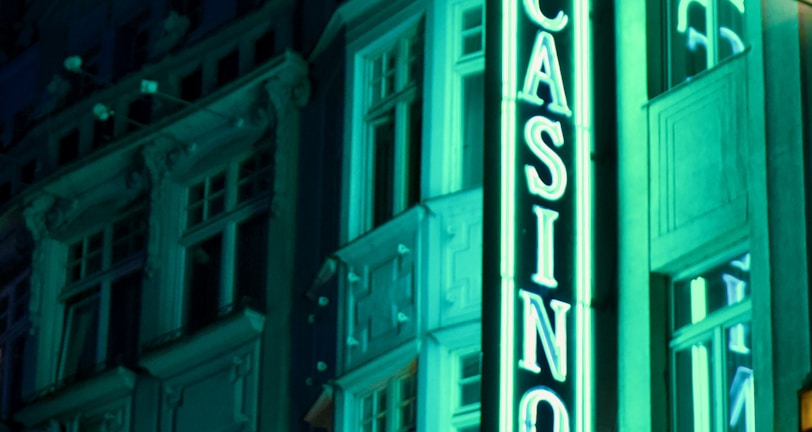 A vibrant neon sign displaying the word 'CASINO' is prominently illuminated against the facade of a building, showcasing intricate architectural details and a classic design. The sign emits a bright teal glow, casting light on the surrounding area and creating a lively atmosphere at night.